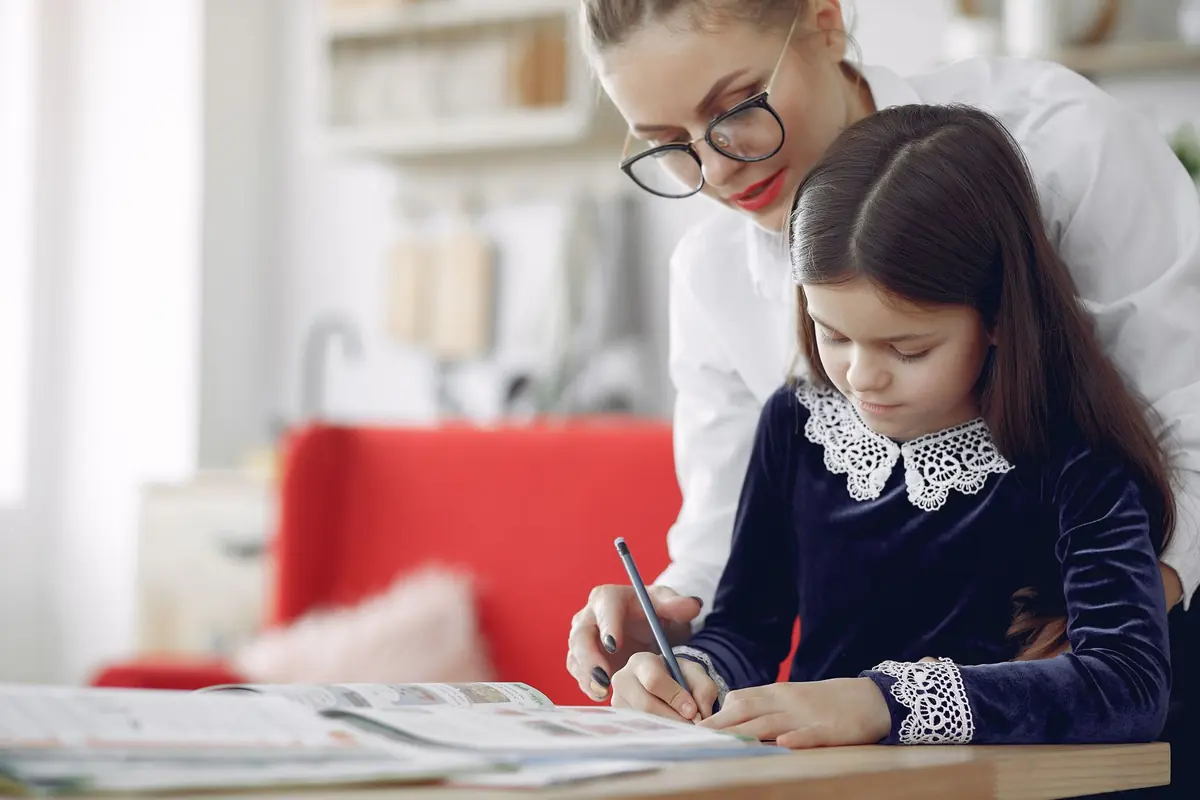 A child writing at a table with an adult helping, in a bright room